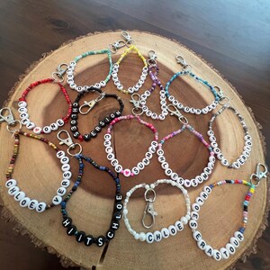 May include: A collection of beaded keychains with white letter beads spelling out names and phrases like "Chloe's", "Summer", and "Hits". The keychains feature various colors of beads and silver-tone clasps, displayed on a wooden surface.