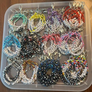 May include: A collection of beaded keychains in a clear plastic container. The keychains feature various colors of beads, with white letter beads spelling out names and words like "Summer", "Chloe", and "Forever". Each keychain has a silver clasp.