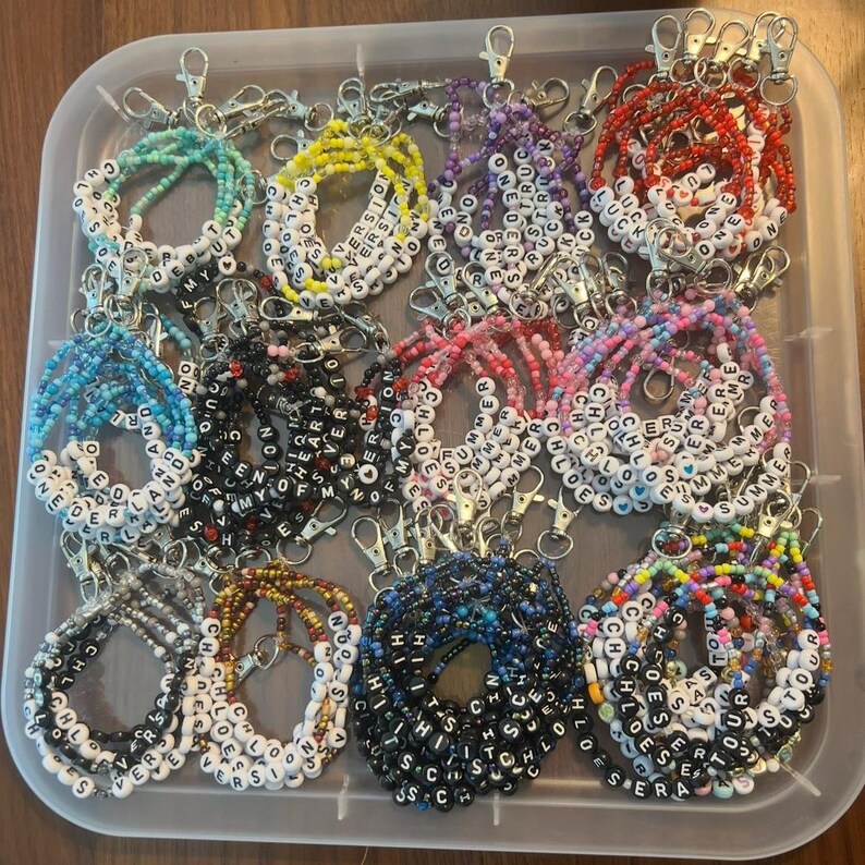 May include: A collection of beaded keychains in a clear plastic container. The keychains feature various colors of beads, with white letter beads spelling out names and words like "Summer", "Chloe", and "Forever". Each keychain has a silver clasp.