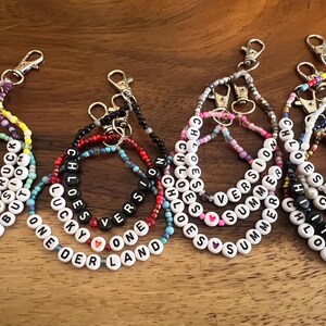 May include: A collection of beaded keychains with white letter beads spelling out names and phrases like "Chloe," "Summer," and "One Version." The keychains feature various colors of beads and silver-tone clasps.
