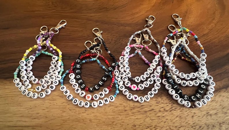 May include: A collection of beaded keychains with white letter beads spelling out names and phrases like "Chloe," "Summer," and "One Version." The keychains feature various colors of beads and silver-tone clasps.