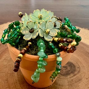 Puede incluir: Una pequeña maceta de terracota llena de musgo verde y adornada con adornos en forma de flor verde claro con ribetes dorados. Hilos de cuentas verdes y marrones caen en cascada de la maceta, creando un arreglo floral decorativo.