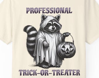 Op de afbeelding: Crèmekleurig T-shirt met een wasbeer verkleed als spook met een pompoen lantaarn. De tekst "PROFESSIONAL TRICK-OR-TREATER" staat boven en onder de wasbeer illustratie in paars.