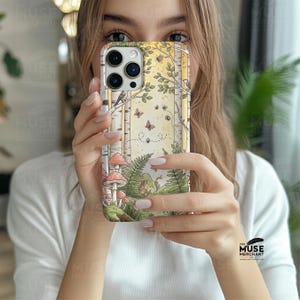 Op de afbeelding: Een telefoonhoesje met een bosillustratie, inclusief berken, paddenstoelen, vlinders en varens. Het ontwerp maakt gebruik van een aquarel-stijl met groene, gele en bruine tinten. De hoes wordt voor een persoon gehouden.