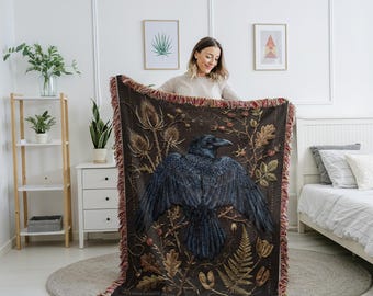 May include: A dark brown throw blanket featuring a detailed illustration of a raven with outstretched wings. The blanket has a fringed edge and botanical elements in shades of gold and brown. The raven is depicted in shades of black and blue.
