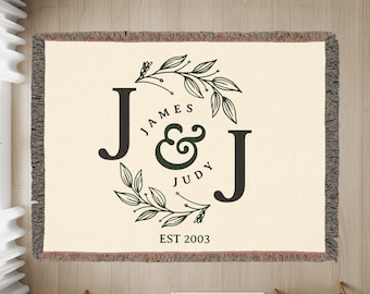 May include: Cream-colored woven throw blanket with a decorative design. The design features the letters "J" on either side of the names "JAMES & JUDY" and the year "EST 2003" in a circular floral wreath. The blanket has a fringed edge.