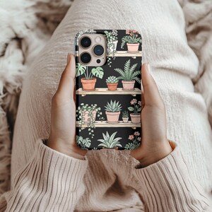 Puede incluir: Una funda de tel&eacute;fono negra con un dise&ntilde;o de plantas en macetas sobre estantes de madera. Las plantas est&aacute;n en macetas de terracota, con follaje verde y peque&ntilde;as flores. La funda est&aacute; sujeta en las manos de una persona.