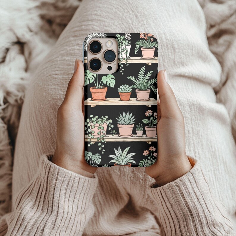 Puede incluir: Una funda de tel&eacute;fono negra con un dise&ntilde;o de plantas en macetas sobre estantes de madera. Las plantas est&aacute;n en macetas de terracota, con follaje verde y peque&ntilde;as flores. La funda est&aacute; sujeta en las manos de una persona.
