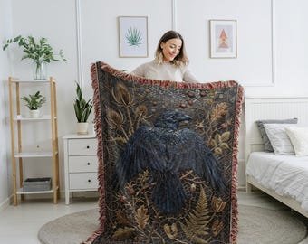 May include: A woven throw blanket featuring a detailed illustration of a raven with outstretched wings. The blanket has a dark background with botanical elements in shades of brown, green, and red. The edges are fringed.