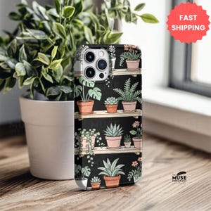 Puede incluir: Funda de tel&eacute;fono negra con un dise&ntilde;o bot&aacute;nico de plantas en macetas sobre estantes de madera. La funda tiene un acabado mate y un recorte para la c&aacute;mara. El logotipo de The Muse Merchant es visible en la parte inferior derecha.