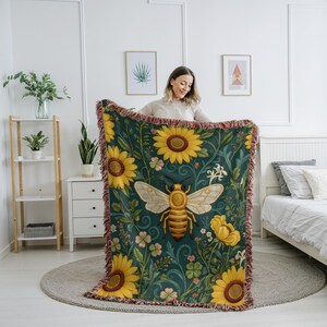 May include: A decorative throw blanket with a bee and floral design. The blanket features a large bee with sunflowers and other flowers on a dark teal background. The edges are fringed with red and brown threads. The blanket's design includes yellow, white, and green accents.