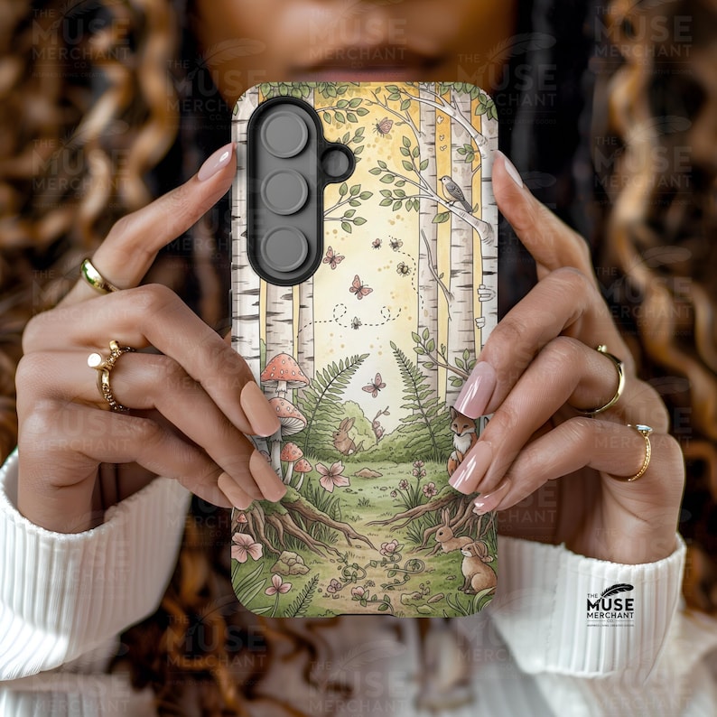 Op de afbeelding: Een telefoonhoesje met een bosillustratie, met berken, vlinders en bosdieren. Het ontwerp bevat paddenstoelen. De hoes heeft een zwarte cameramodule en wordt door een persoon vastgehouden.