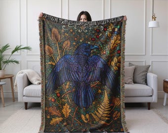 May include: A woven tapestry throw blanket featuring a detailed illustration of a raven with outstretched wings. The design incorporates elements of nature, including leaves, berries, and thistle, in shades of blue, green, and gold. The blanket has a fringed edge.