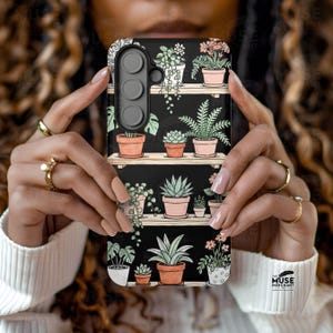 Puede incluir: Funda de tel&eacute;fono negra con dise&ntilde;o bot&aacute;nico. Presenta plantas en macetas sobre estantes de madera, con plantas verdes y flores rosas en macetas de terracota sobre un fondo oscuro. El logotipo de The Muse Merchant es visible.