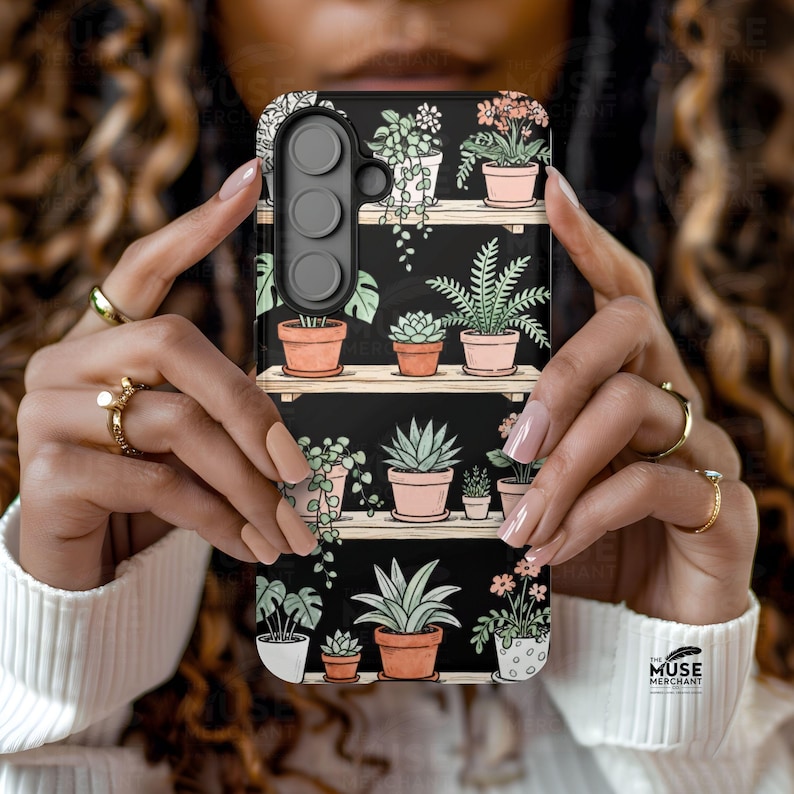 Puede incluir: Funda de tel&eacute;fono negra con dise&ntilde;o bot&aacute;nico. Presenta plantas en macetas sobre estantes de madera, con plantas verdes y flores rosas en macetas de terracota sobre un fondo oscuro. El logotipo de The Muse Merchant es visible.