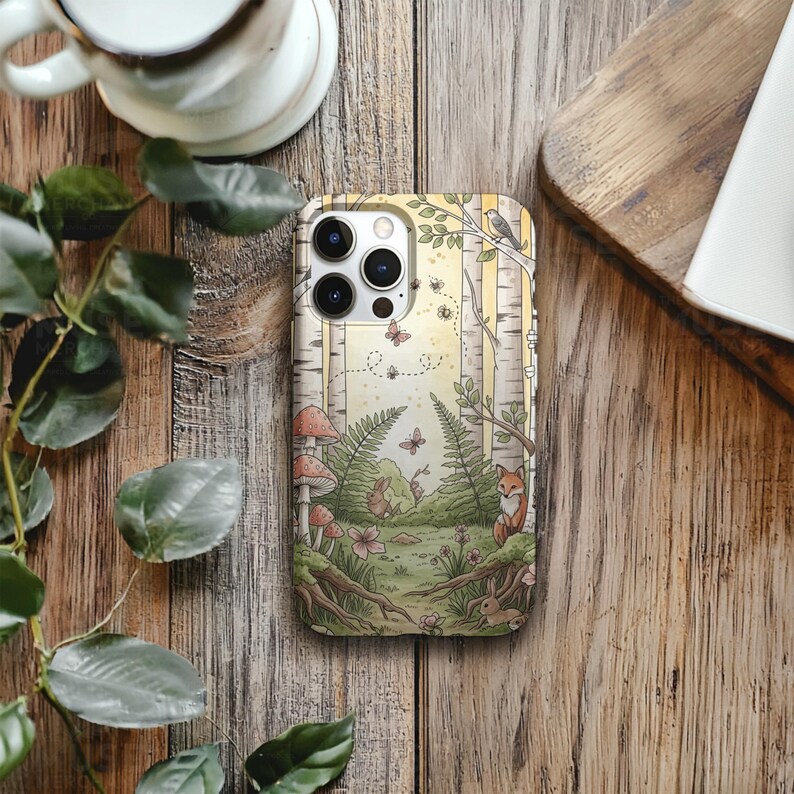 Op de afbeelding: Telefoonhoesje met een bosillustratie, inclusief berken, paddenstoelen en een vos. Het ontwerp toont vlinders, varens en een konijn in gedempte tinten. Gepresenteerd op een houten oppervlak.