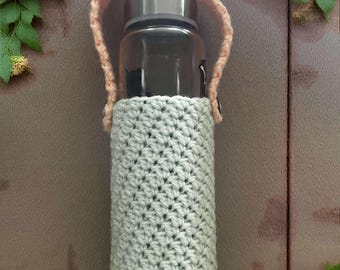 May include: A clear glass water bottle with a silver lid, encased in a light gray crocheted cozy. The cozy has a matching peach-colored handle. The bottle is surrounded by pink and peach flowers.