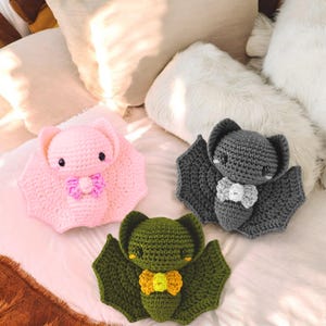May include: Three handmade crochet bats in pink, gray, and green. Each bat has large wings, small eyes, and a bow tie. The bats are displayed on a white surface with pillows in the background.