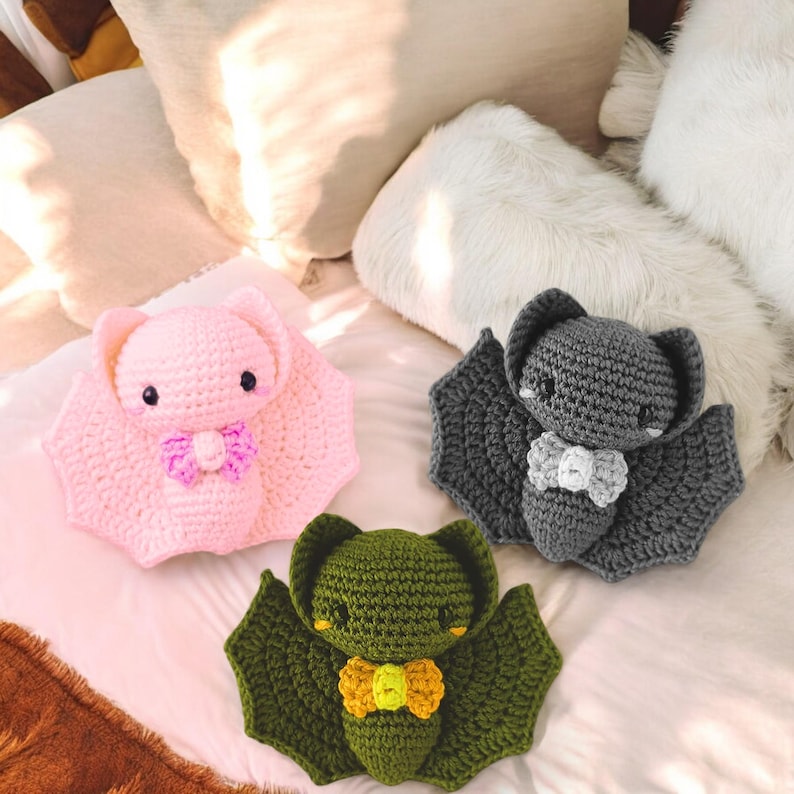 May include: Three handmade crochet bats in pink, gray, and green. Each bat has large wings, small eyes, and a bow tie. The bats are displayed on a white surface with pillows in the background.