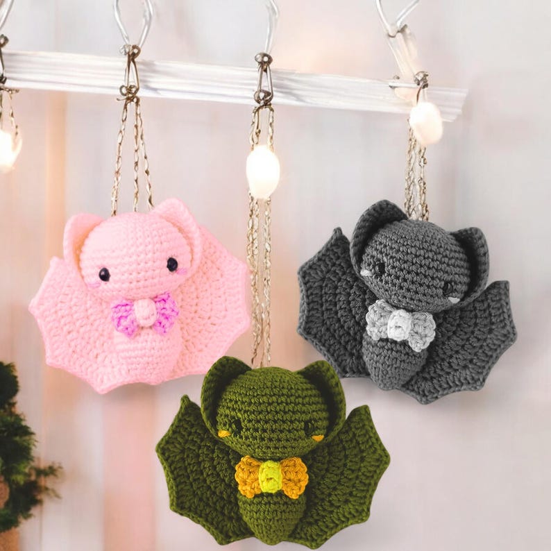 May include: Three crocheted bat ornaments in pink, gray, and green, each with a bow tie. The bats are hanging from a white shelf with string lights. The pink bat has a pink bow tie, the gray bat has a white bow tie, and the green bat has a yellow bow tie.