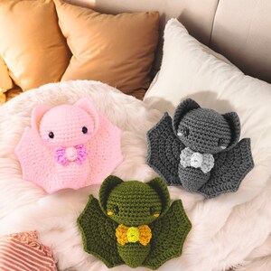 May include: Three crocheted bat plushies in pink, gray, and green, each with a bow tie. The pink bat has a purple bow, the gray bat has a white bow, and the green bat has a yellow bow. The bats are on a white, fluffy surface.