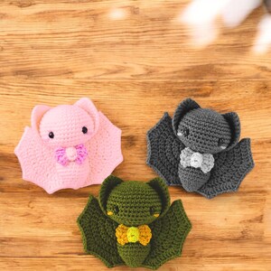 May include: Three handmade crochet bats in pink, gray, and green, each with a bow tie. The bats have large wings and small, round eyes. The bats are displayed on a wooden surface.