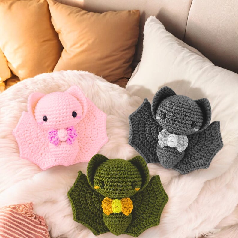 May include: Three crocheted bat plushies in pink, gray, and green, each with a bow tie. The pink bat has a purple bow, the gray bat has a white bow, and the green bat has a yellow bow. The bats are on a white, fluffy surface.