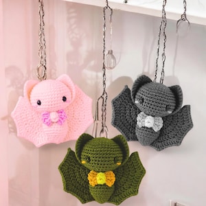 May include: Three crocheted bat ornaments in pink, gray, and green, each with a bow tie. The bats are hanging from silver chains. The pink bat has a purple bow tie, the gray bat has a white bow tie, and the green bat has a yellow bow tie.