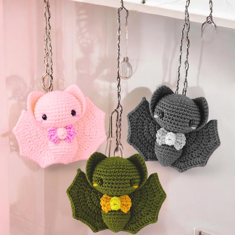 May include: Three crocheted bat ornaments in pink, gray, and green, each with a bow tie. The bats are hanging from silver chains. The pink bat has a purple bow tie, the gray bat has a white bow tie, and the green bat has a yellow bow tie.