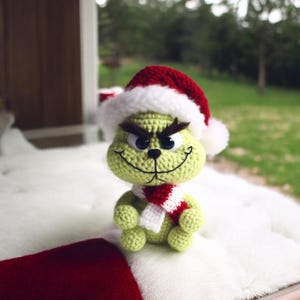 May include: A crocheted Grinch doll wearing a red and white Santa hat and a red and white striped scarf. The Grinch has a green body, black eyes, and a mischievous smile. The doll is sitting on a white surface.