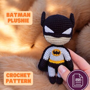 May include: A handmade Batman plushie with a black cowl, white eyes, and a gray and yellow costume. The text "BATMAN PLUSHIE" and "CROCHET PATTERN" are displayed. The plushie is held in a hand.