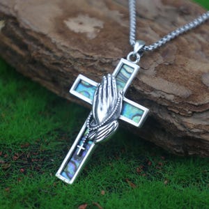 Puede incluir: Collar con colgante de cruz plateada. La cruz presenta un diseño detallado de manos rezando y un rosario. La cruz está incrustada con concha de abulón iridiscente. El collar se muestra sobre una superficie de madera natural.