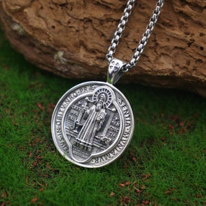 May include: A silver pendant necklace with a circular medallion depicting a religious figure and text. The pendant is suspended from a silver chain. The background is a mix of green and brown.