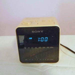 May include: A beige Sony clock radio with a black front panel featuring a digital display showing 1:00 AM in blue light. The device includes an analog radio tuner scale for FM and AM frequencies, with control knobs located on the top surface.