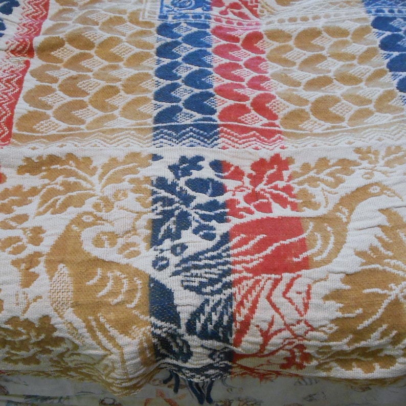 May include: A vintage textile with a repeating pattern of birds, leaves, and geometric shapes. The color palette includes shades of gold, red, blue, and white. The fabric appears to be woven with a textured surface.