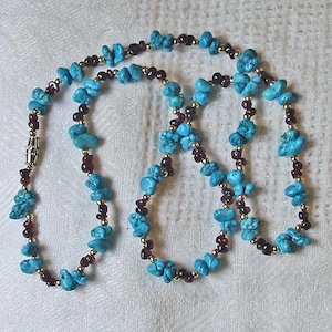 May include: A beaded necklace featuring alternating raw turquoise nuggets and small dark red garnet beads, accented with tiny gold-toned spacer beads. The necklace is finished with a cylindrical metal clasp closure, creating a colorful pattern.
