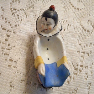 May include: A vintage ceramic clown figurine, featuring a white coat with black buttons, blue pants, and yellow cuffs. The clown has a black hat with a red pom-pom and a ruffled collar. The figurine is designed to hold small items.