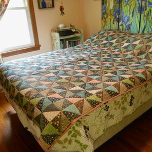 May include: A bed quilt featuring a geometric pattern of triangles in shades of green, brown, peach, and blue. The quilt is constructed from various patterned fabrics and is finished with a solid peach-colored binding along the edges.