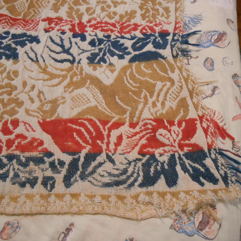 May include: A vintage textile featuring a repeating pattern of tan deer, red flowers, and blue foliage on an off-white background. The textile has a fringed edge and a textured appearance, suggesting it may be a blanket or throw.