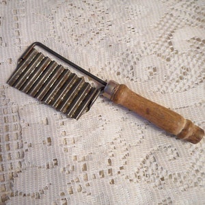 May include: A vintage metal and wood potato chipper with a wavy blade. The chipper has a wooden handle and a metal blade with a wavy edge.