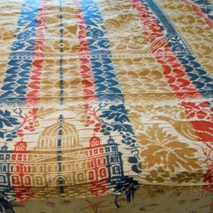 May include: Close-up of a vintage textile with a repeating pattern. The design features alternating vertical stripes in blue, red, and tan, with floral and architectural motifs. The textile has a textured appearance and a faded color palette.