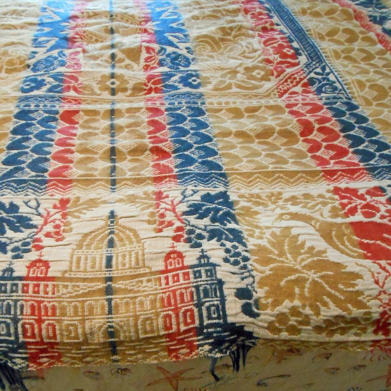 May include: Close-up of a vintage textile with a repeating pattern. The design features alternating vertical stripes in blue, red, and tan, with floral and architectural motifs. The textile has a textured appearance and a faded color palette.