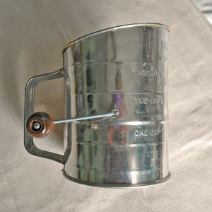 May include: A vintage metal flour sifter with a handle and a wooden knob. The sifter has measurements for one, two, and three cups. The handle is attached to the side of the sifter, and the wooden knob is on the side.