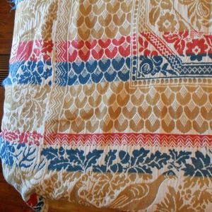 May include: Cream-colored vintage textile with intricate patterns. Features repeating heart shapes in tan, red, and blue stripes. Floral and bird motifs are also present, creating a detailed and textured design. The textile has a fringed edge.