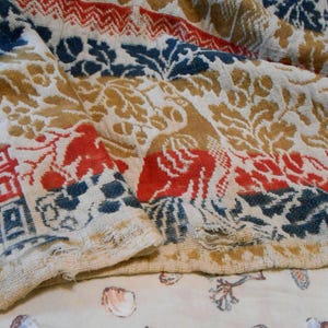 May include: Close-up of a vintage textile with a repeating pattern. The design features red, blue, and gold floral and geometric motifs on an off-white background. The fabric appears worn, with visible fraying along the edges, suggesting age and use.