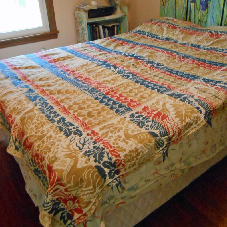 May include: A vintage bedspread with a repeating pattern of deer, birds, and floral designs in shades of tan, red, and blue. The bedspread is rectangular and covers the bed. The bedspread is made of a soft, possibly quilted material.
