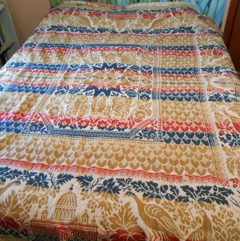 May include: A vintage bedspread with a detailed pattern in red, blue, and gold. The design includes floral, geometric, and bird motifs. The bedspread is made of a soft, possibly woven, fabric.