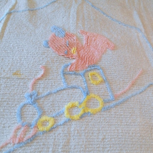 May include: A cream-colored towel with a textured surface. The towel features an embroidered design of a pink and yellow creature riding a blue and yellow vehicle. The design is outlined in blue and pink yarn.