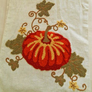 May include: A light beige fabric runner featuring a central punch needle pumpkin design in vibrant orange and yellow yarn. The pumpkin is surrounded by embroidered olive green leaves, brown curling vines, and small yellow star-shaped flowers.