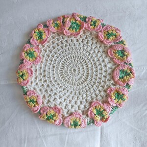 May include: A round, off-white crocheted doily featuring a central open-work pattern and a decorative border composed of multiple three-dimensional floral motifs in shades of pink, yellow, and green, arranged in a circular formation.